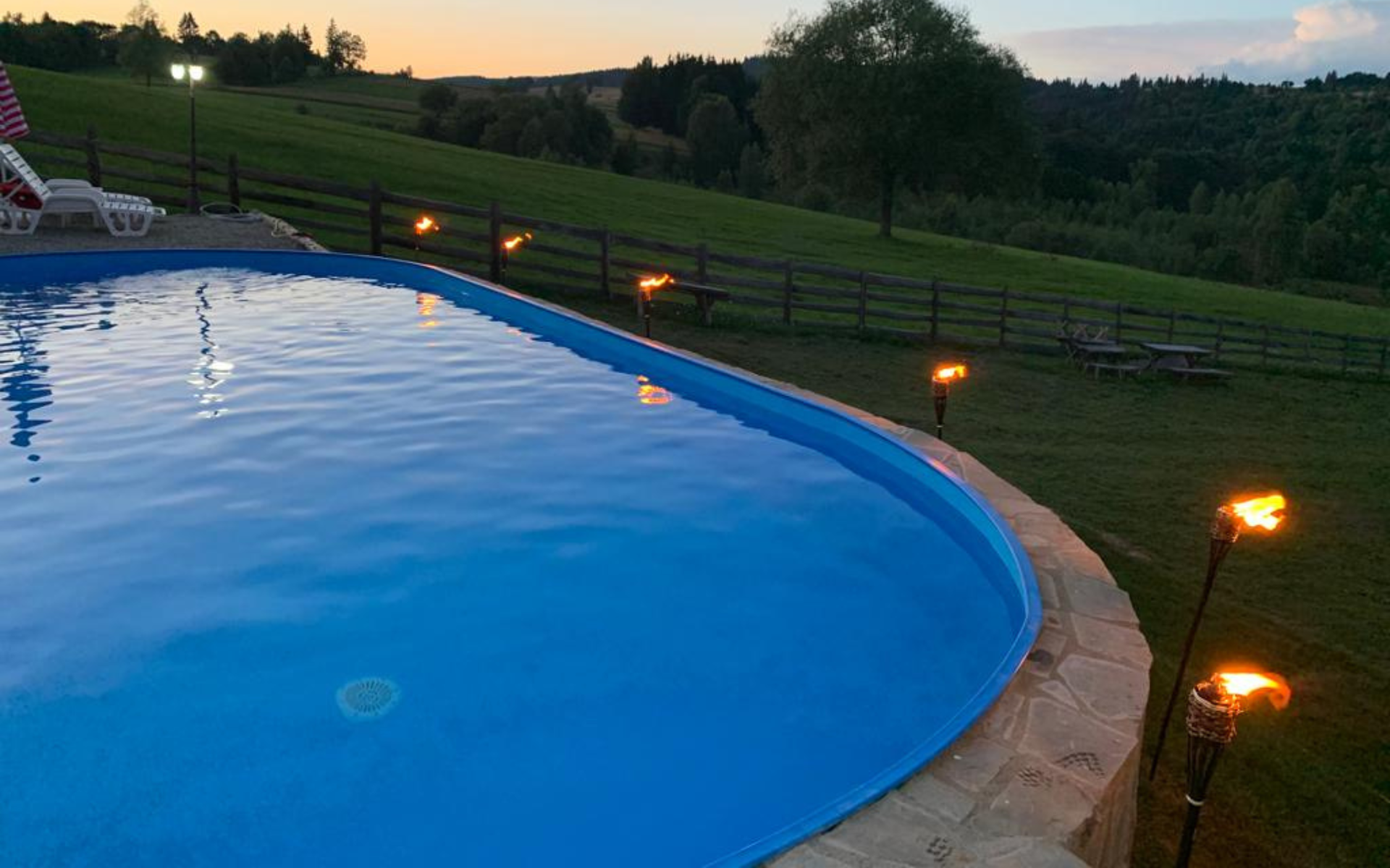 Piscină exterioară la Casa Brădet, iluminată cu torțe la apus, vedere spre dealuri verzi